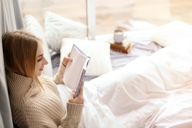 Beautiful young woman in knitted sweater sitting and reading book near window at home. Winter atmosphere