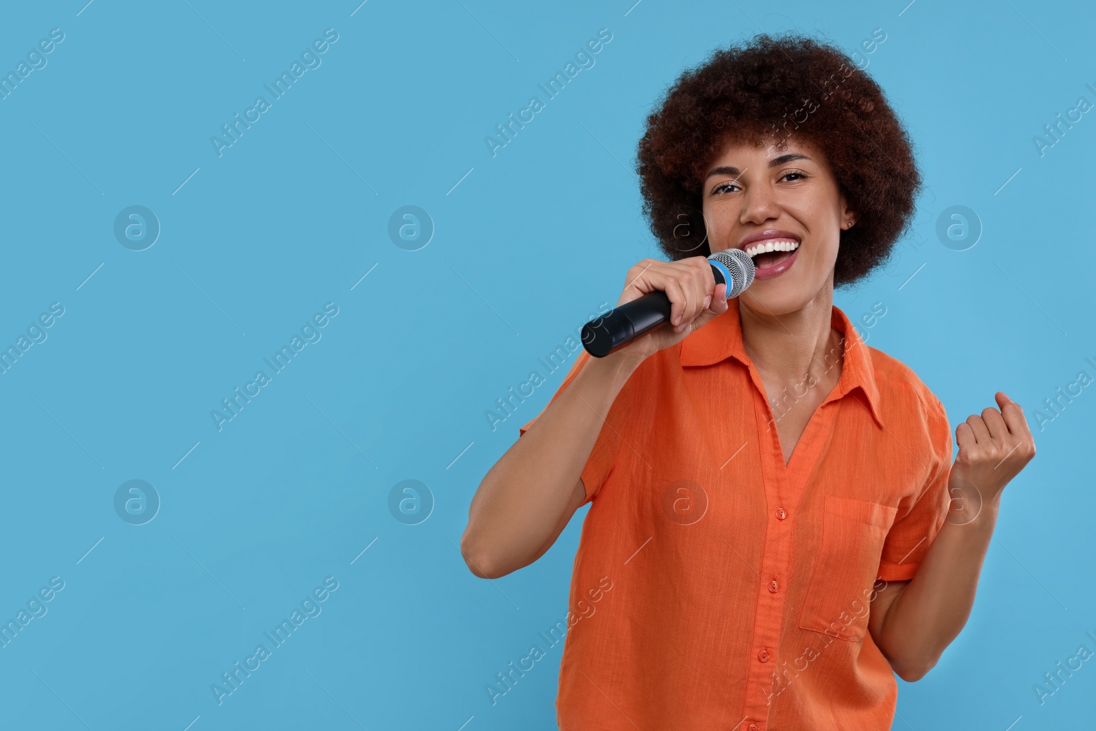 Photo of Curly young woman with microphone singing on light blue background, space for text