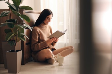 Photo of Woman with cupdrink and book sitting on warm floor at home. Heating system