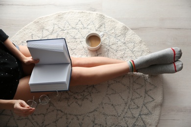 Photo of Young woman with cup of coffee reading book on floor at home, top view