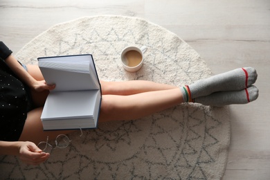 Young woman with cup of coffee reading book on floor at home, top view