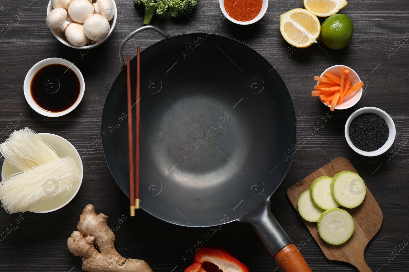 Photo of Empty iron wok and chopsticks surrounded by ingredients on dark grey wooden table, flat lay