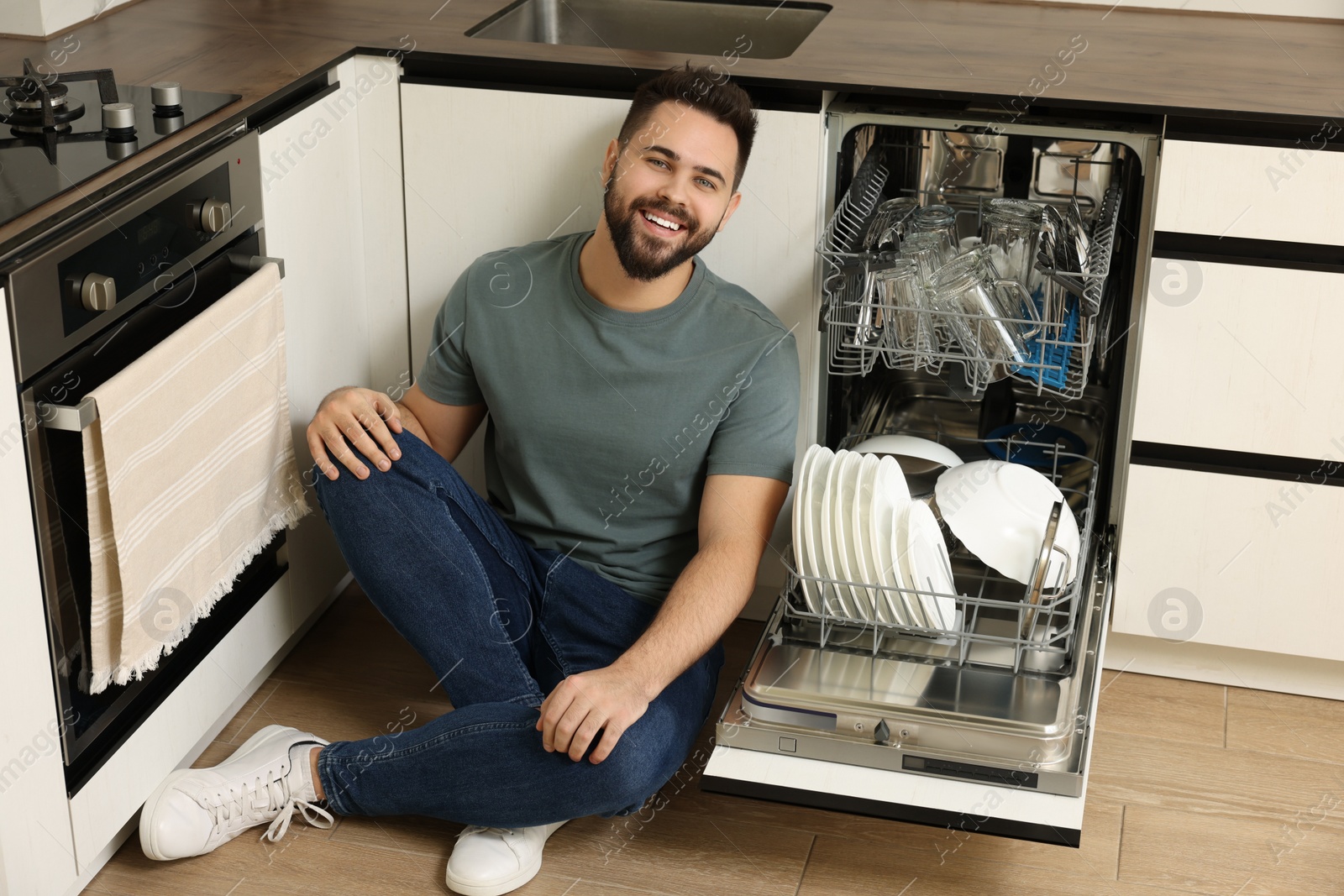 Photo of Smiling man sitting near open dishwasher in kitchen