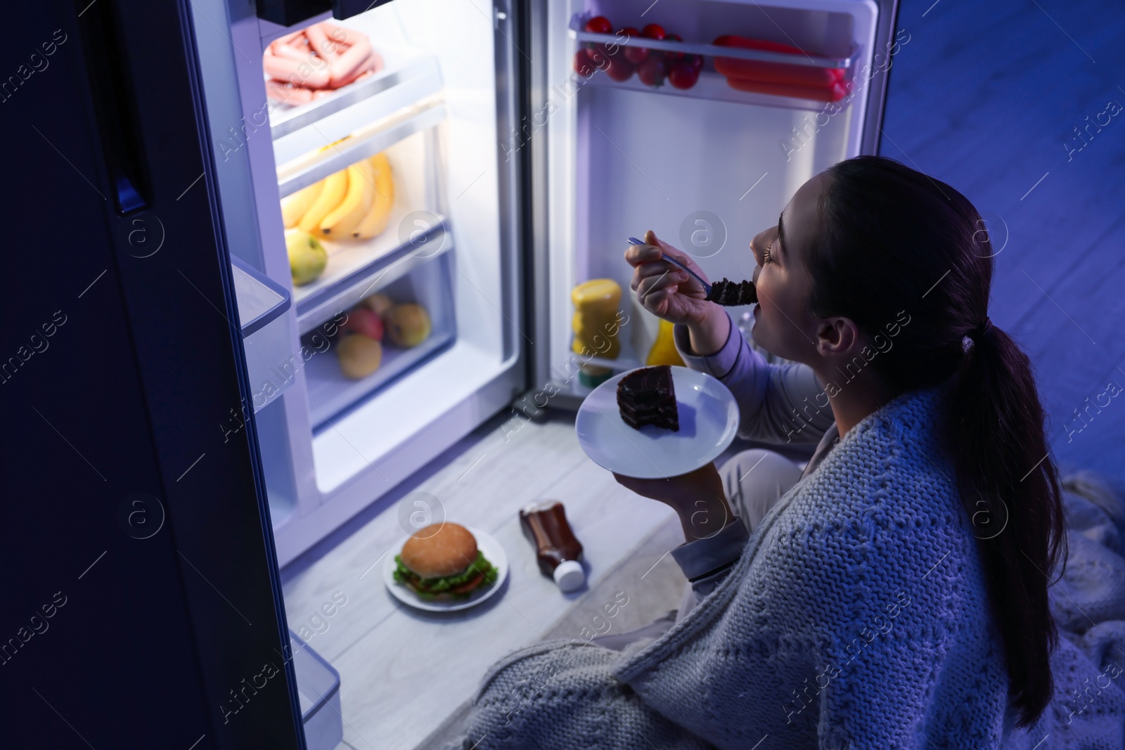 Photo of Young woman eating cake near refrigerator in kitchen at night. Bad habit