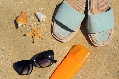 Stylish sunglasses, slippers and bottle of sunblock on sand, flat lay. Beach accessories