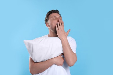 Photo of Sleepy man with pillow yawning on light blue background. Insomnia problem