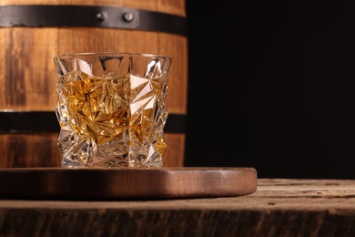 Whiskey with ice cubes in glass and barrel on wooden table against black background, closeup. Space for text