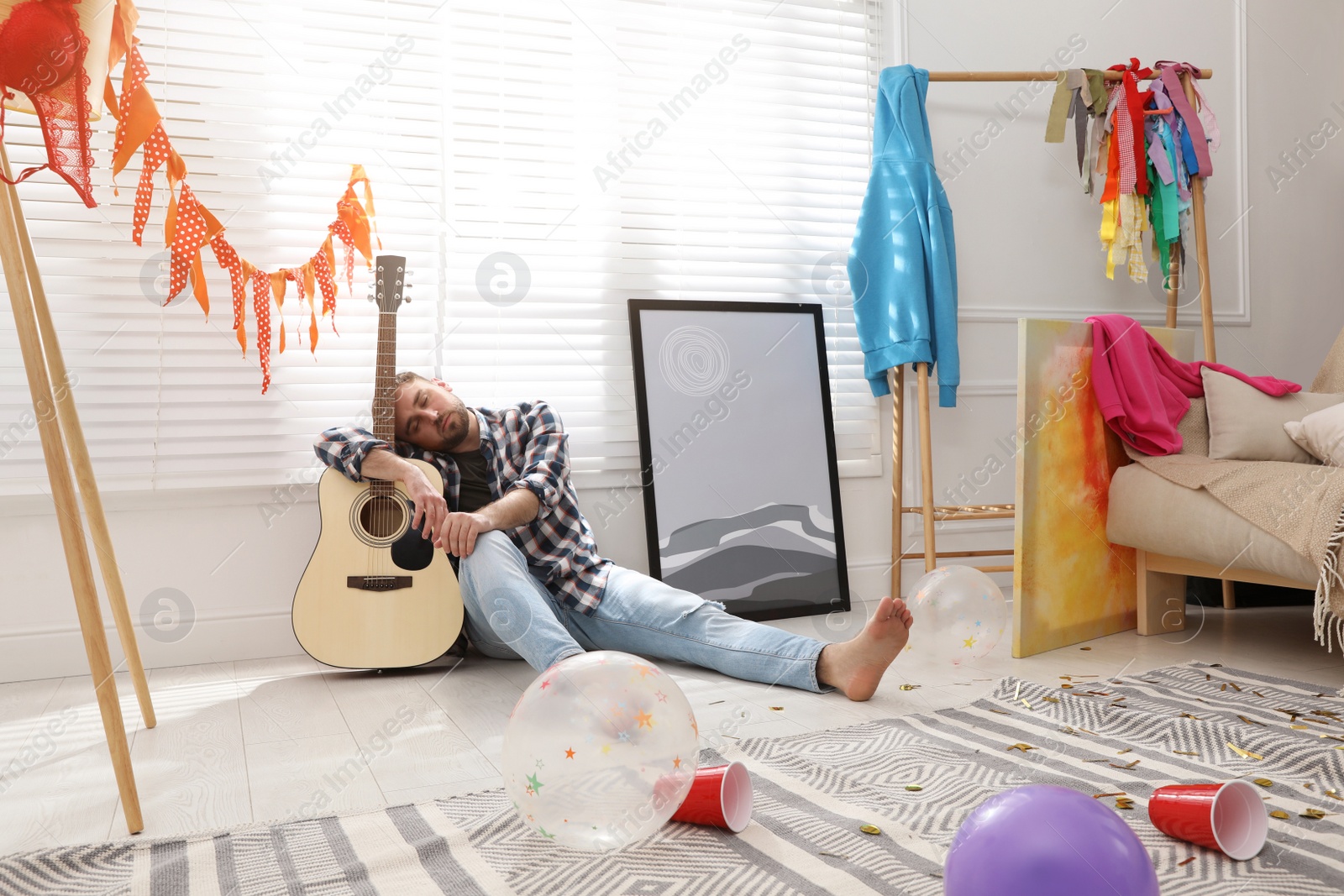 Photo of Young man with guitar sleeping near window in messy room after party