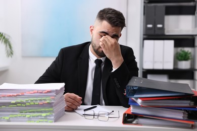 Overwhelmed man sitting at table with stacks of documents and folders in office