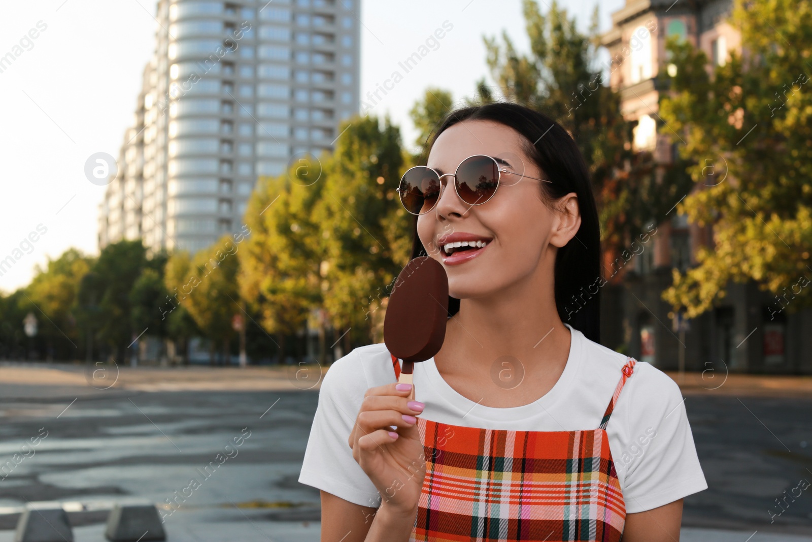 Photo of Beautiful young woman eating ice cream glazed in chocolate on city street