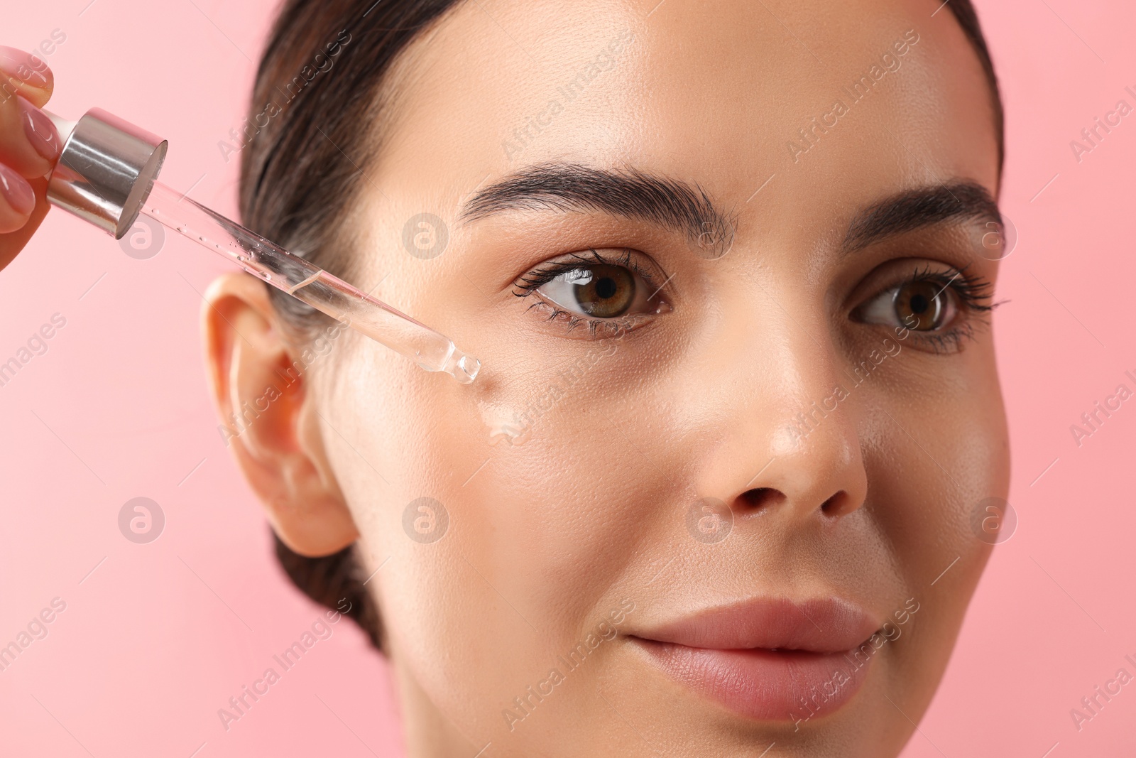 Photo of Beautiful young woman applying serum onto her face on pink background, closeup