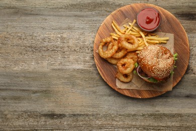 Photo of Top view of serving board with tasty burger, French fries and fried onion rings on wooden table, space for text. Fast food