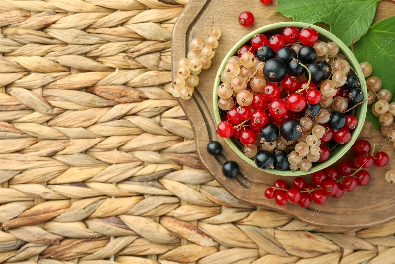 Photo of Different fresh ripe currants and green leaves on wicker surface, flat lay. Space for text