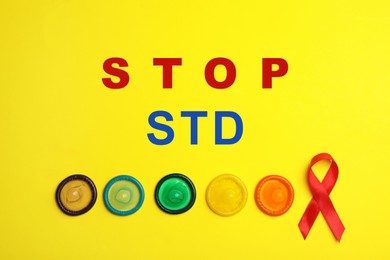 Colorful condoms, red ribbon and text STOP STD on yellow background, flat lay