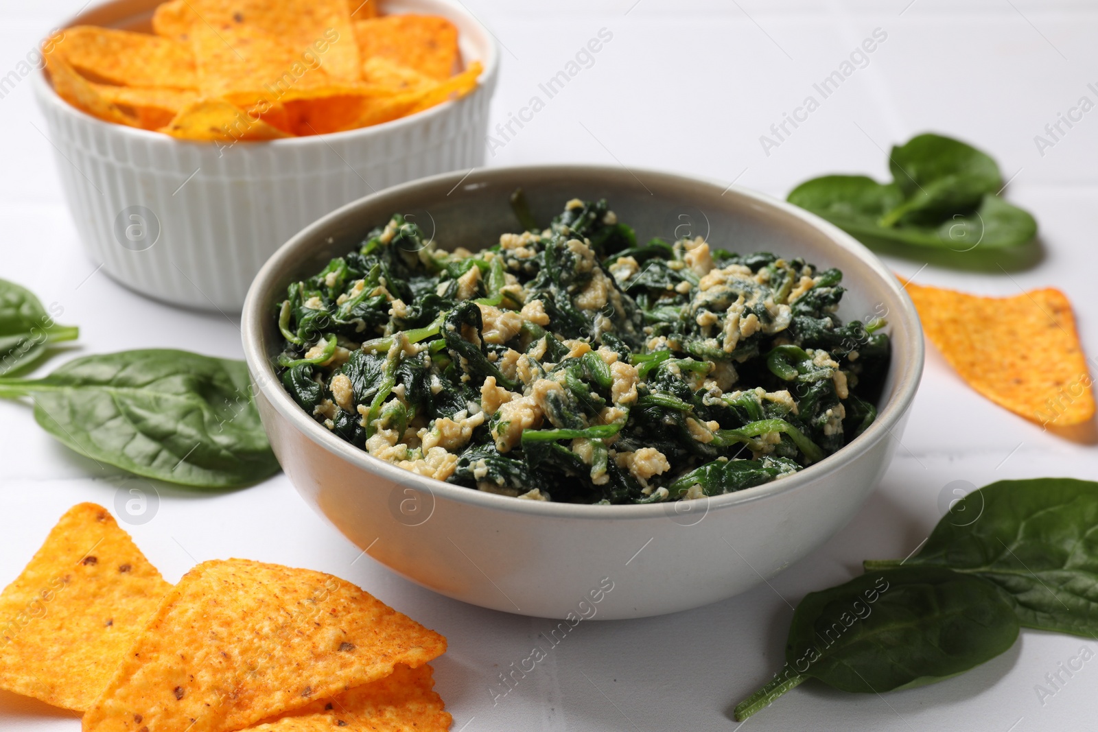 Photo of Tasty spinach dip with egg in bowl and nachos chips on white table, closeup