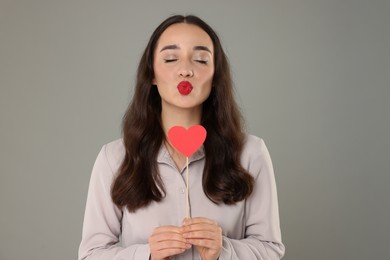 Beautiful young woman with paper heart sending air kiss on grey background