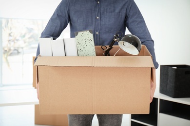 Photo of Young man holding moving box with office stuff indoors, closeup