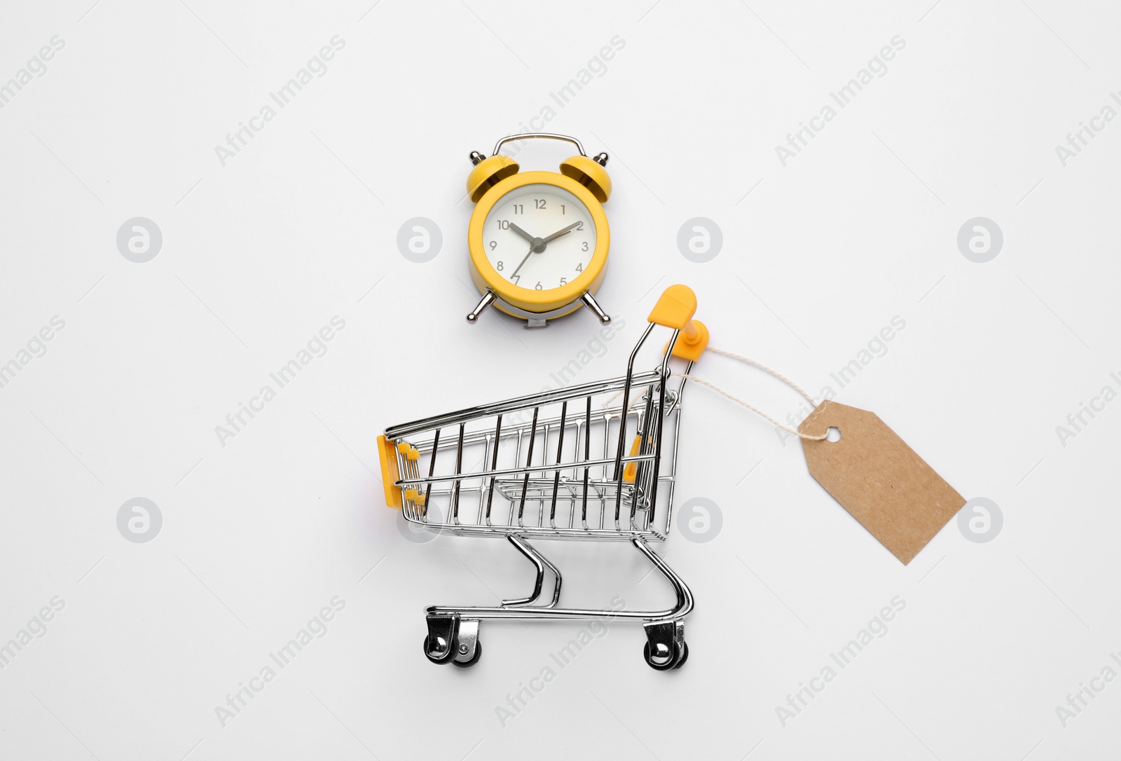 Photo of Shopping cart with tag and alarm clock on white background, top view. Sale concept