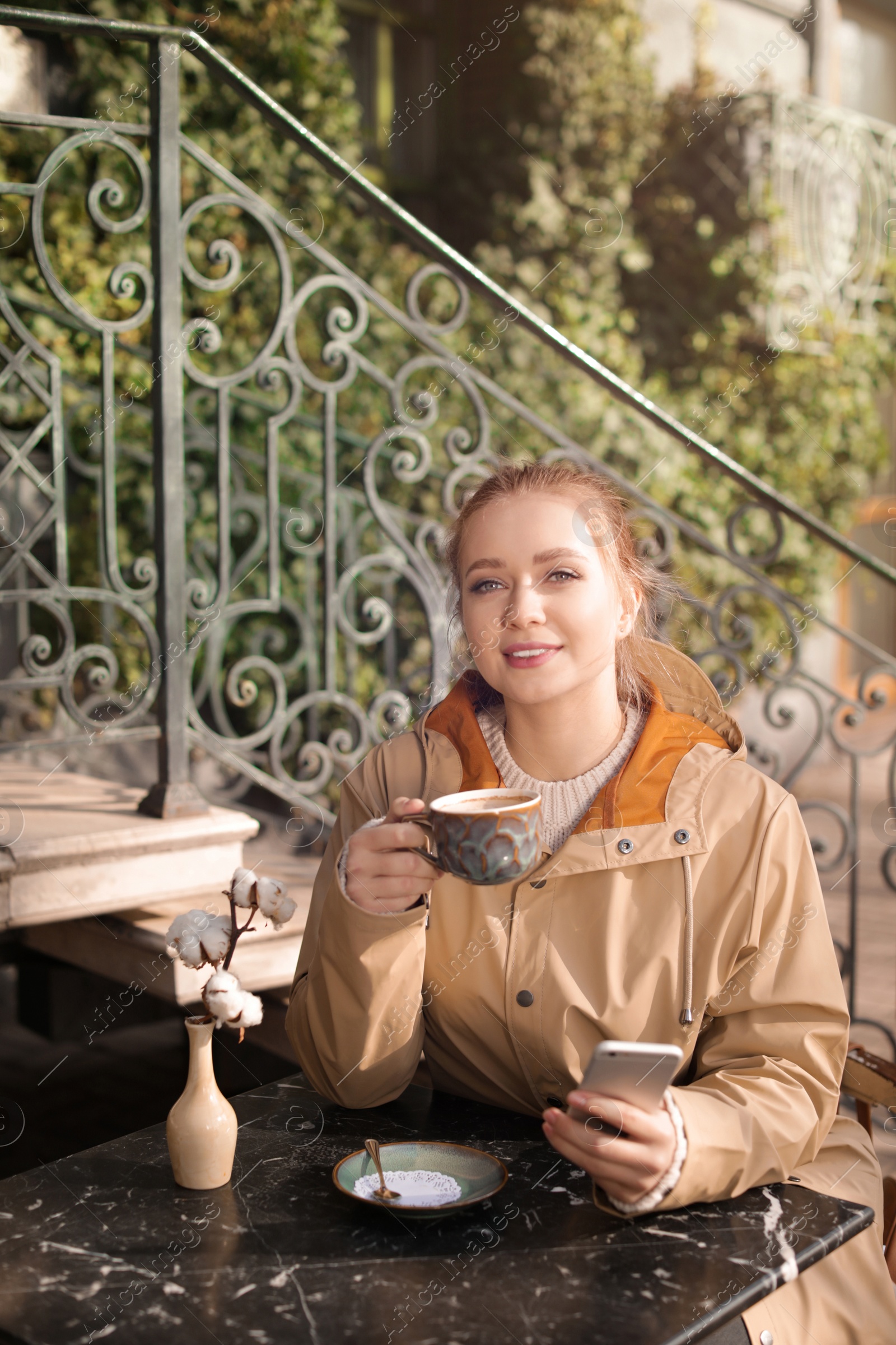 Photo of Young woman enjoying tasty coffee while using mobile phone at table outdoors