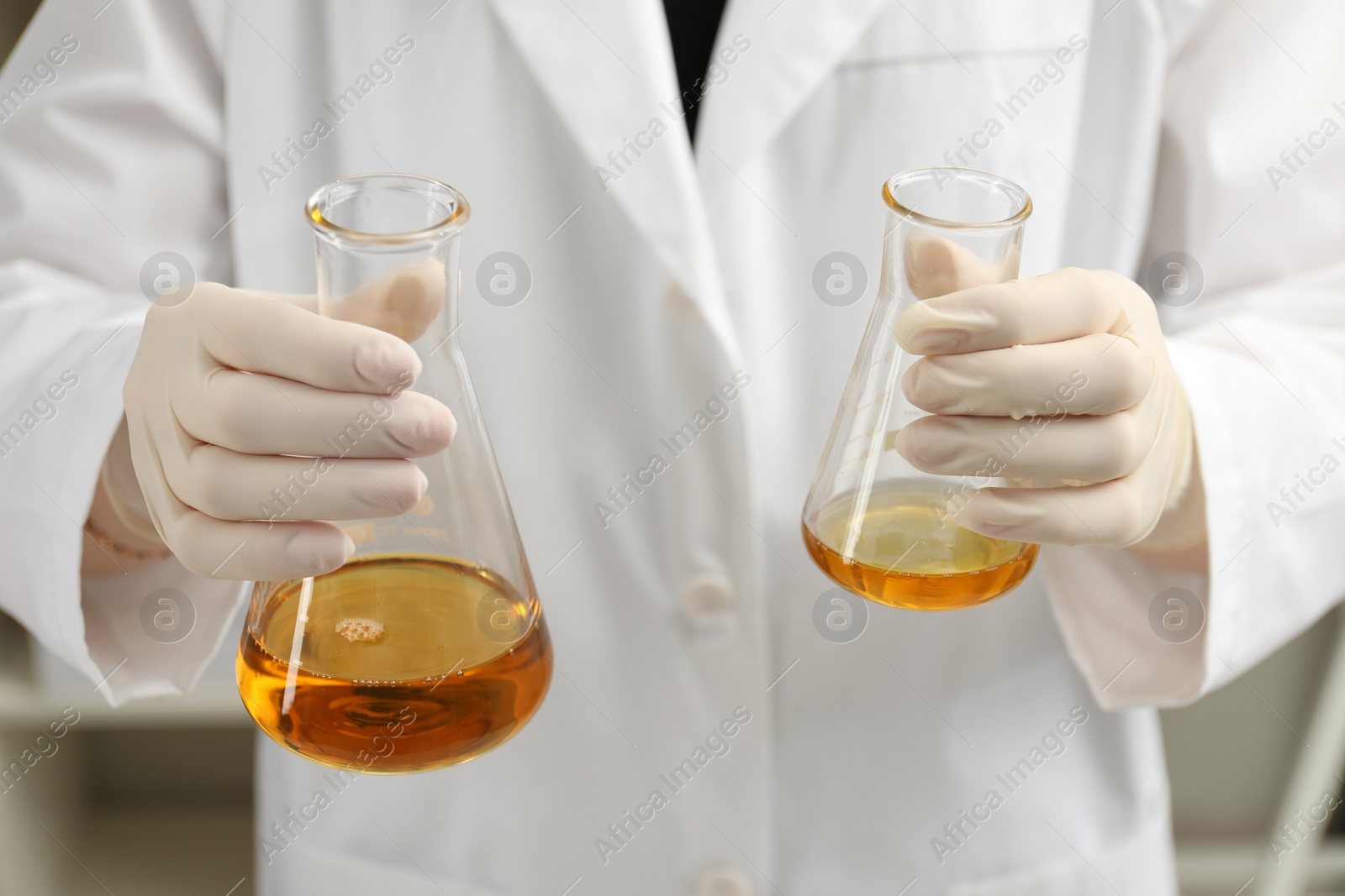 Photo of Laboratory worker holding flasks with orange crude oil, closeup