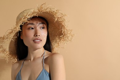 Photo of Beautiful young woman in straw hat with sun protection cream on her face against beige background, space for text