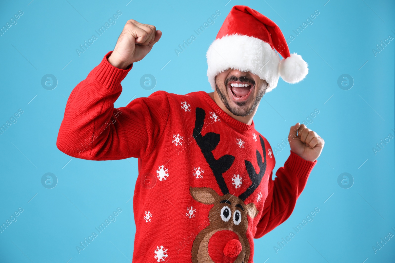 Photo of Emotional young man in Christmas sweater and Santa hat on light blue background