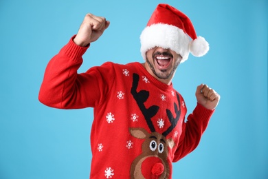 Emotional young man in Christmas sweater and Santa hat on light blue background