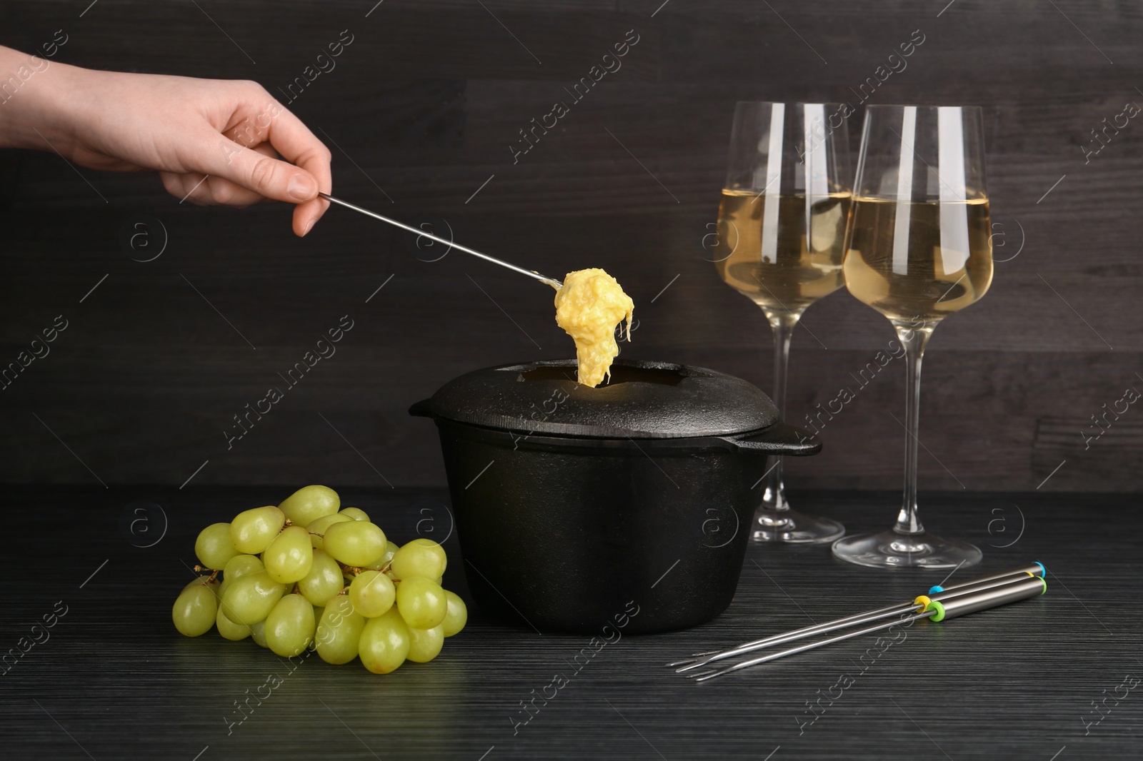 Photo of Woman dipping grape into fondue pot with melted cheese at black wooden table, closeup