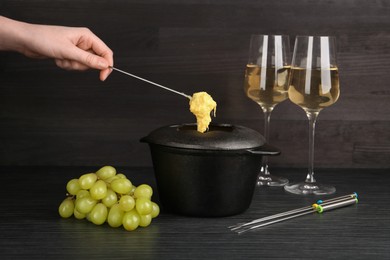 Photo of Woman dipping grape into fondue pot with melted cheese at black wooden table, closeup