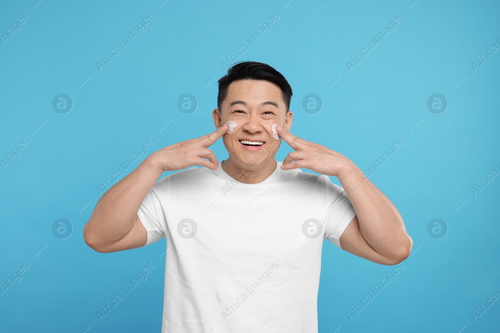 Photo of Happy man applying cream onto his face on light blue background