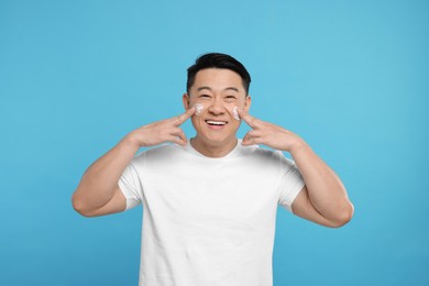 Photo of Happy man applying cream onto his face on light blue background