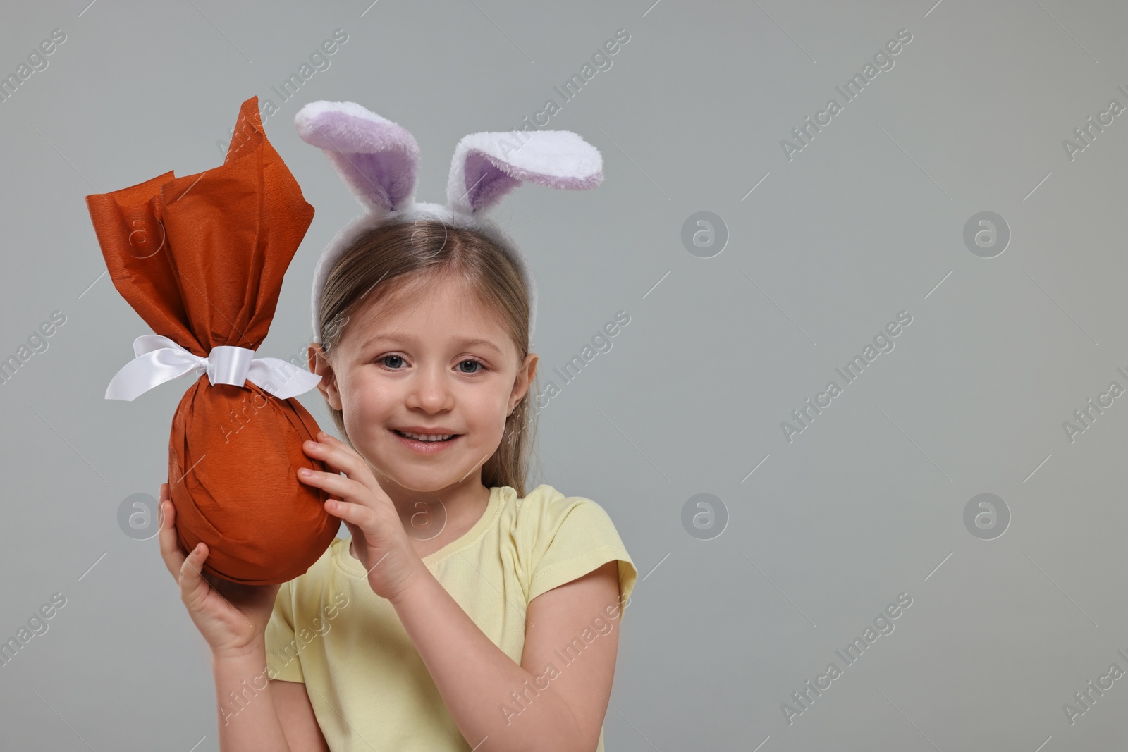 Photo of Easter celebration. Cute girl with bunny ears holding wrapped gift on gray background, space for text
