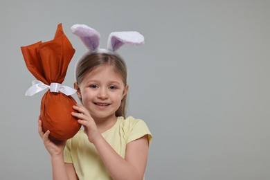 Easter celebration. Cute girl with bunny ears holding wrapped gift on gray background, space for text