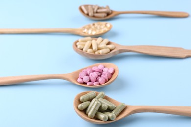 Different vitamin pills in wooden spoons on light blue background, closeup