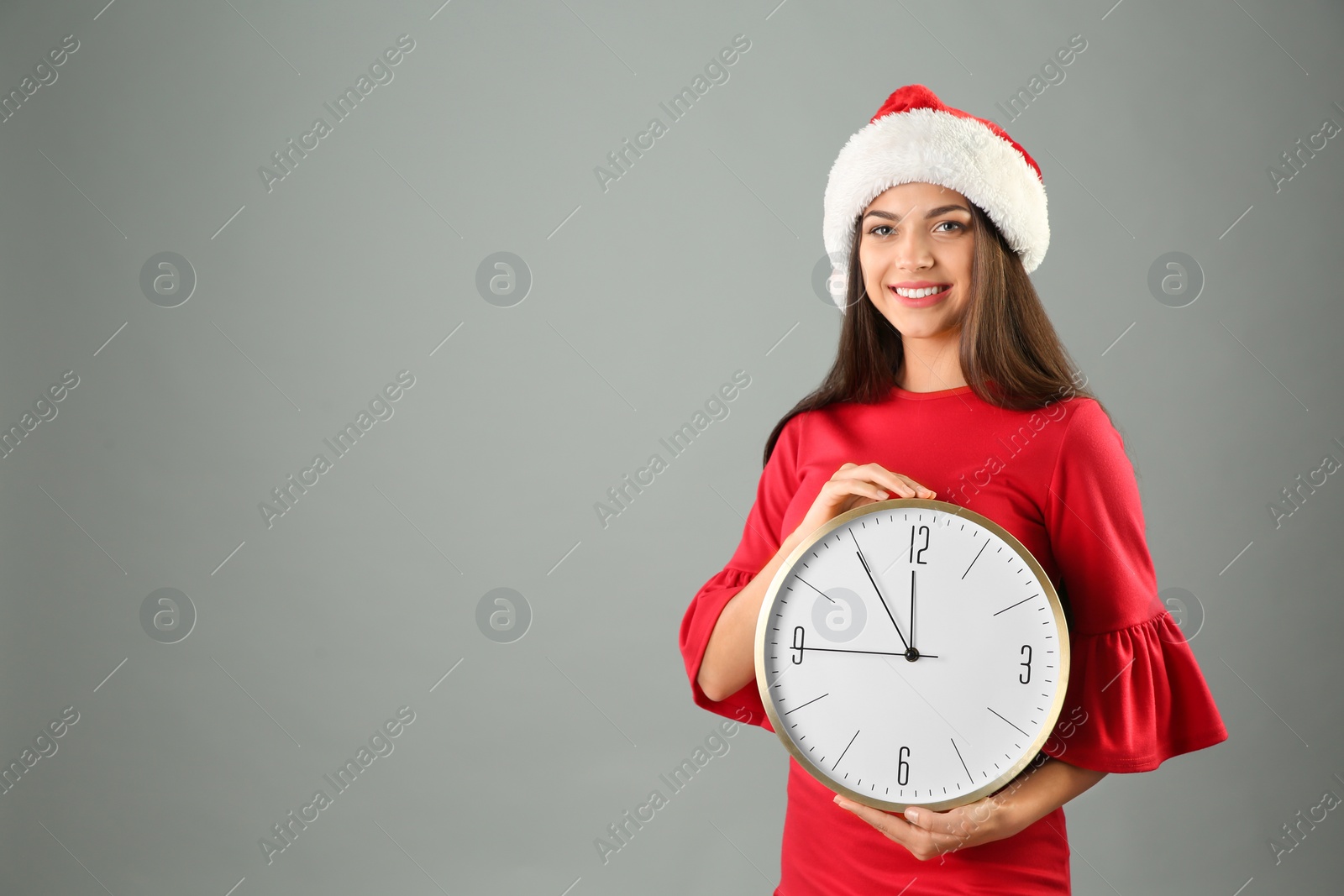 Photo of Young beautiful woman in Santa hat holding big clock on grey background. Christmas celebration