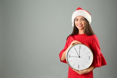 Photo of Young beautiful woman in Santa hat holding big clock on grey background. Christmas celebration