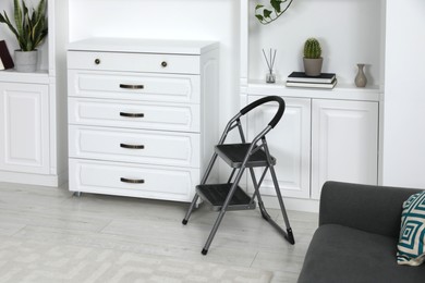 Photo of Metal folding ladder near chest of drawers and shelves with accessories in room
