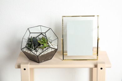 Florarium with succulent plants and photo frame on table near white wall, space for design. Home decor