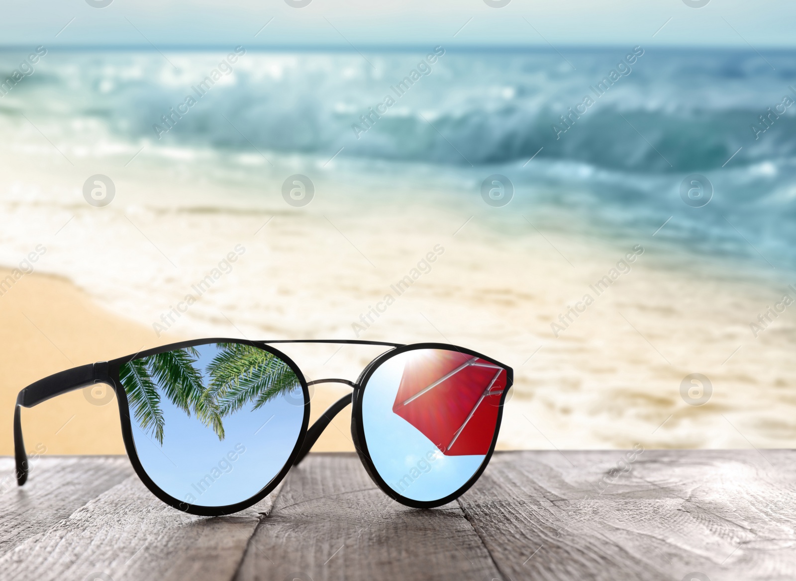 Image of Green palm leaves and sun umbrella mirroring in sunglasses on wooden desk at sandy beach