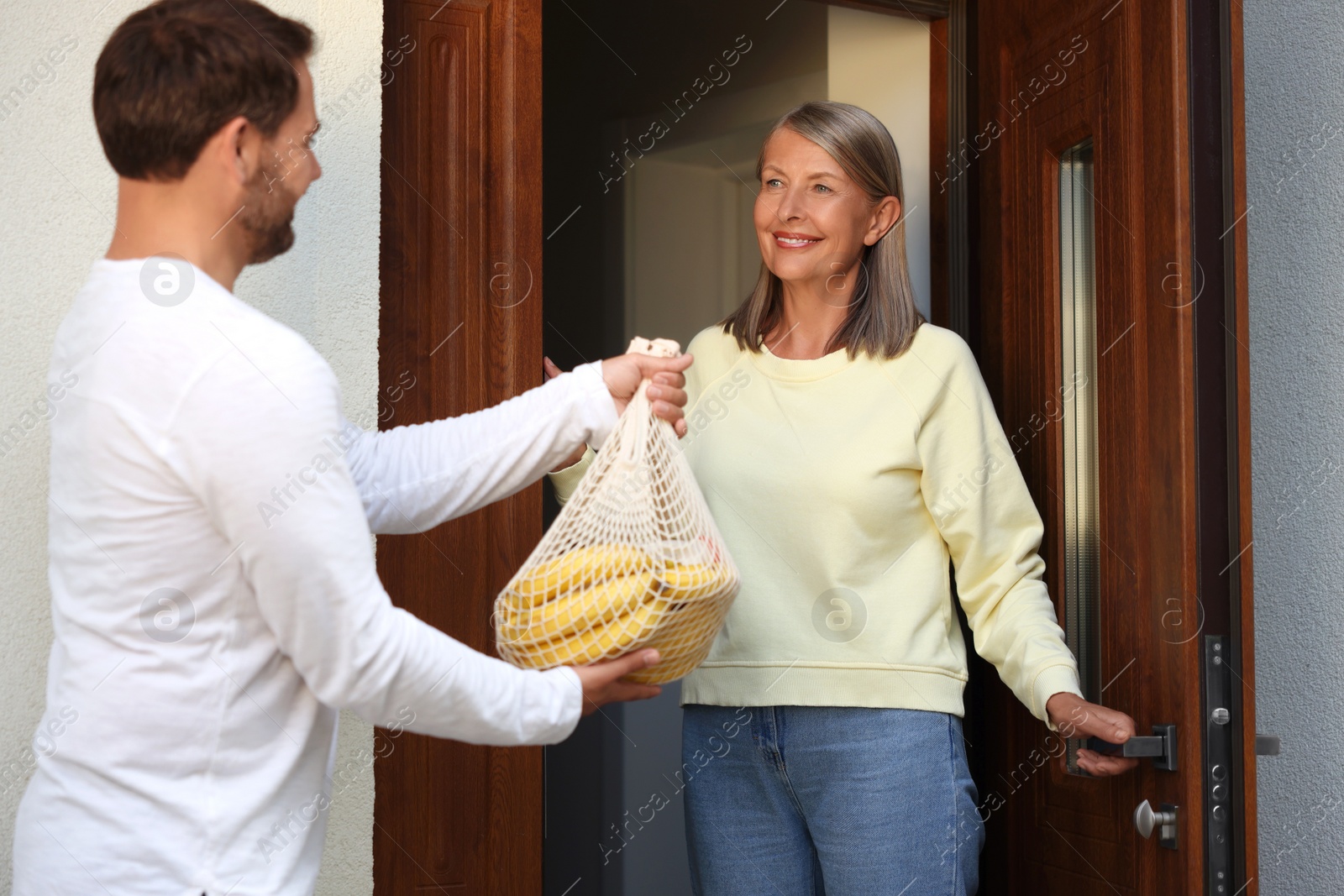 Photo of Helping neighbours. Man with net bag of products visiting senior woman outdoors