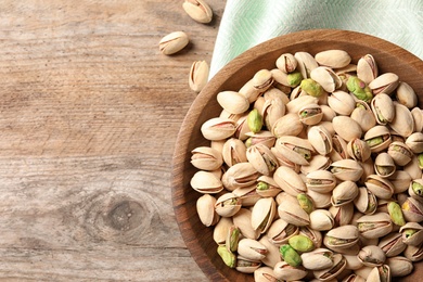 Photo of Organic pistachio nuts in bowl on wooden table, top view. Space for text