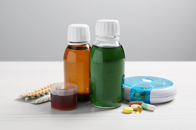 Photo of Bottles of syrup, measuring cup and pills on white table against light grey background. Cold medicine