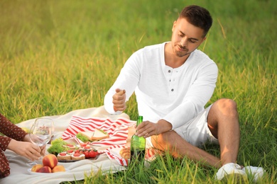 Photo of Young man uncorking wine and his girlfriend in green park. Picnic season