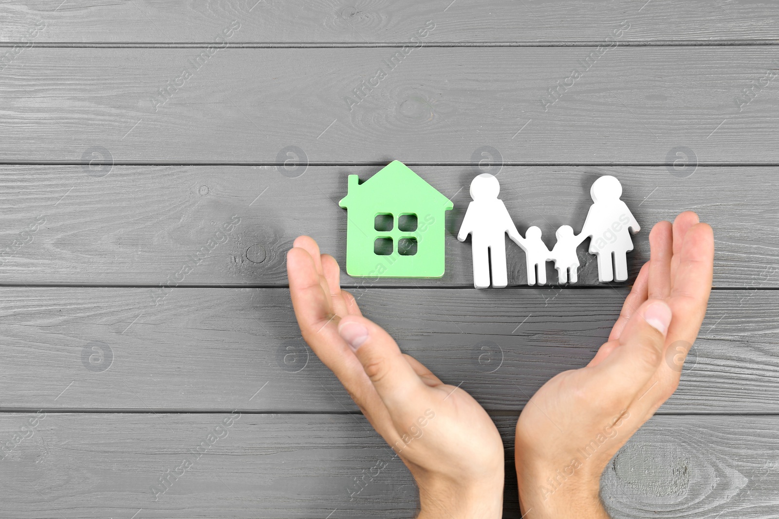 Photo of Young man protecting family figure and house model with his hands on grey wooden background, top view. Space for text