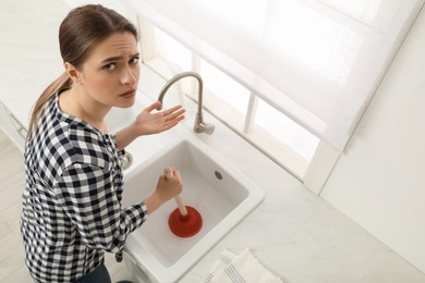 Photo of Upset young woman using plunger to unclog sink drain in kitchen, above view