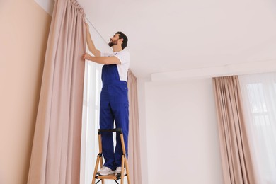 Photo of Worker in uniform hanging window curtain indoors, low angle view