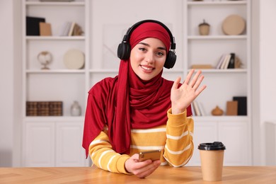 Muslim woman in hijab using smartphone at wooden table in room