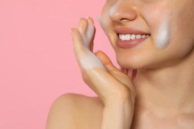 Photo of Young woman washing face with cleansing foam on pink background, closeup and space for text. Skin care cosmetic