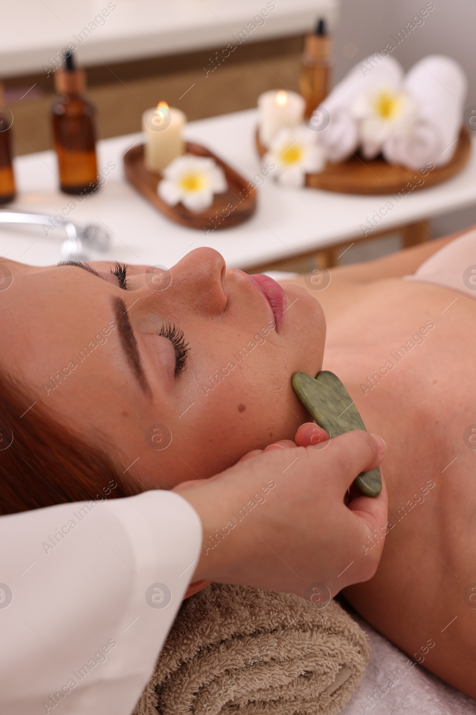 Photo of Young woman receiving facial massage with jade gua sha tool in beauty salon, closeup