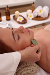Photo of Young woman receiving facial massage with jade gua sha tool in beauty salon, closeup